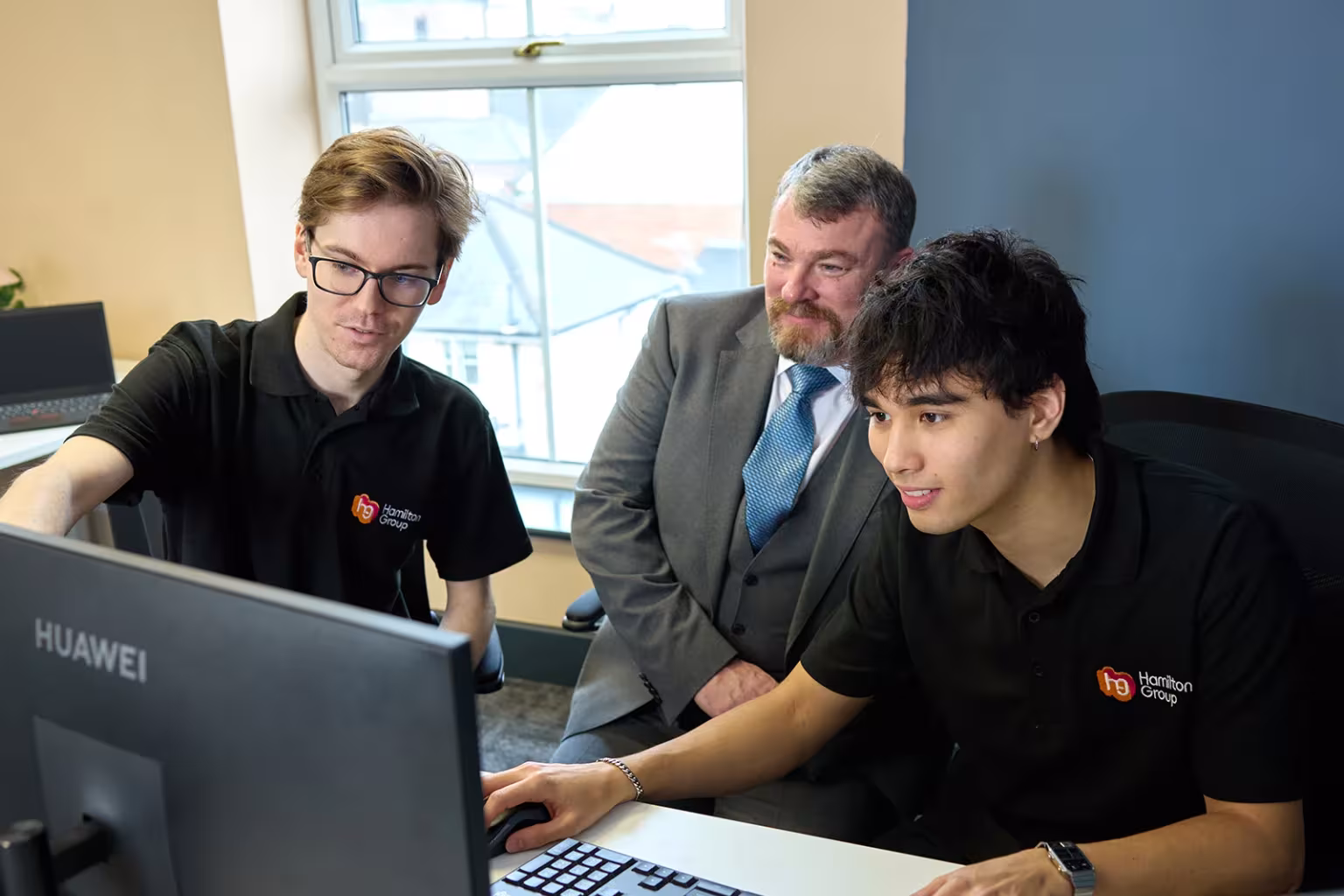 Team collaborating at a computer, with two people in Hamilton Group polo shirts and one in a suit.