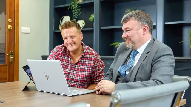 Two men reviewing something on a laptop at a meeting table, smiling during discussion.