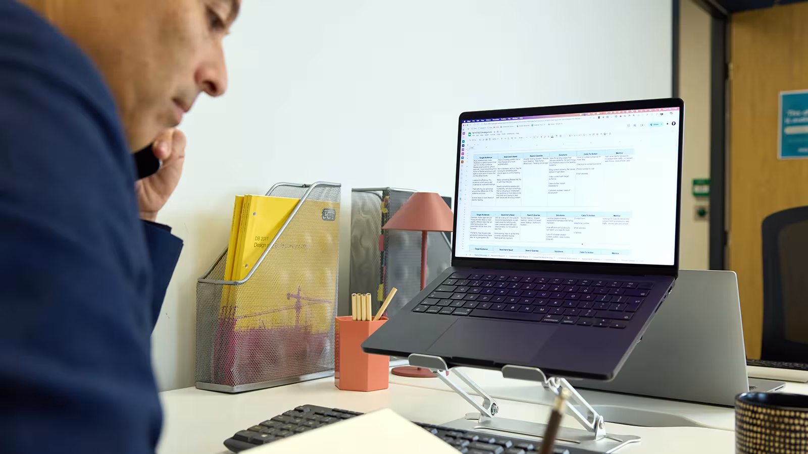 Person on a phone call while taking notes at a desk with a laptop on a stand.
