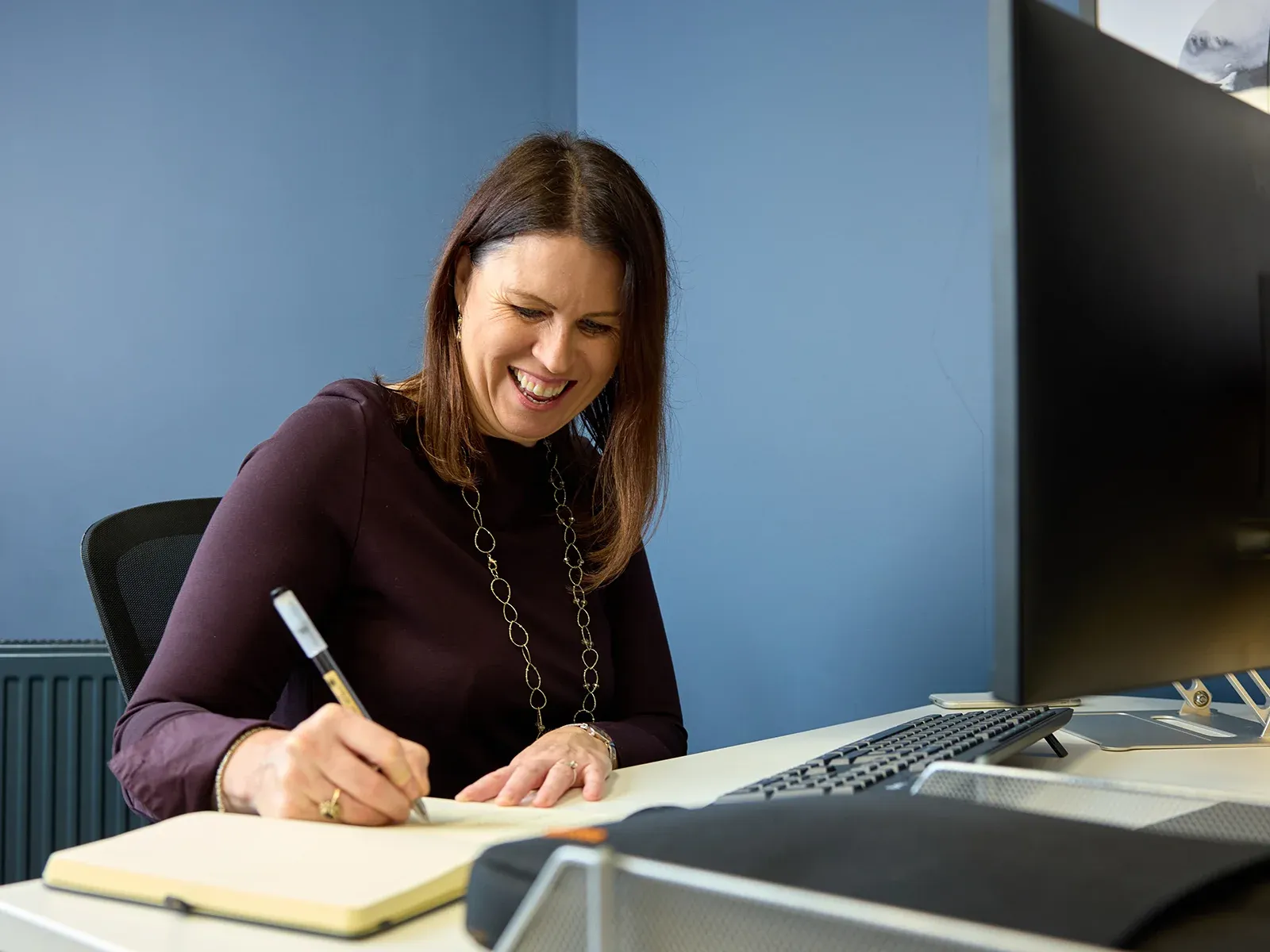 Smiling person writing in a notebook while working at a computer in an office.