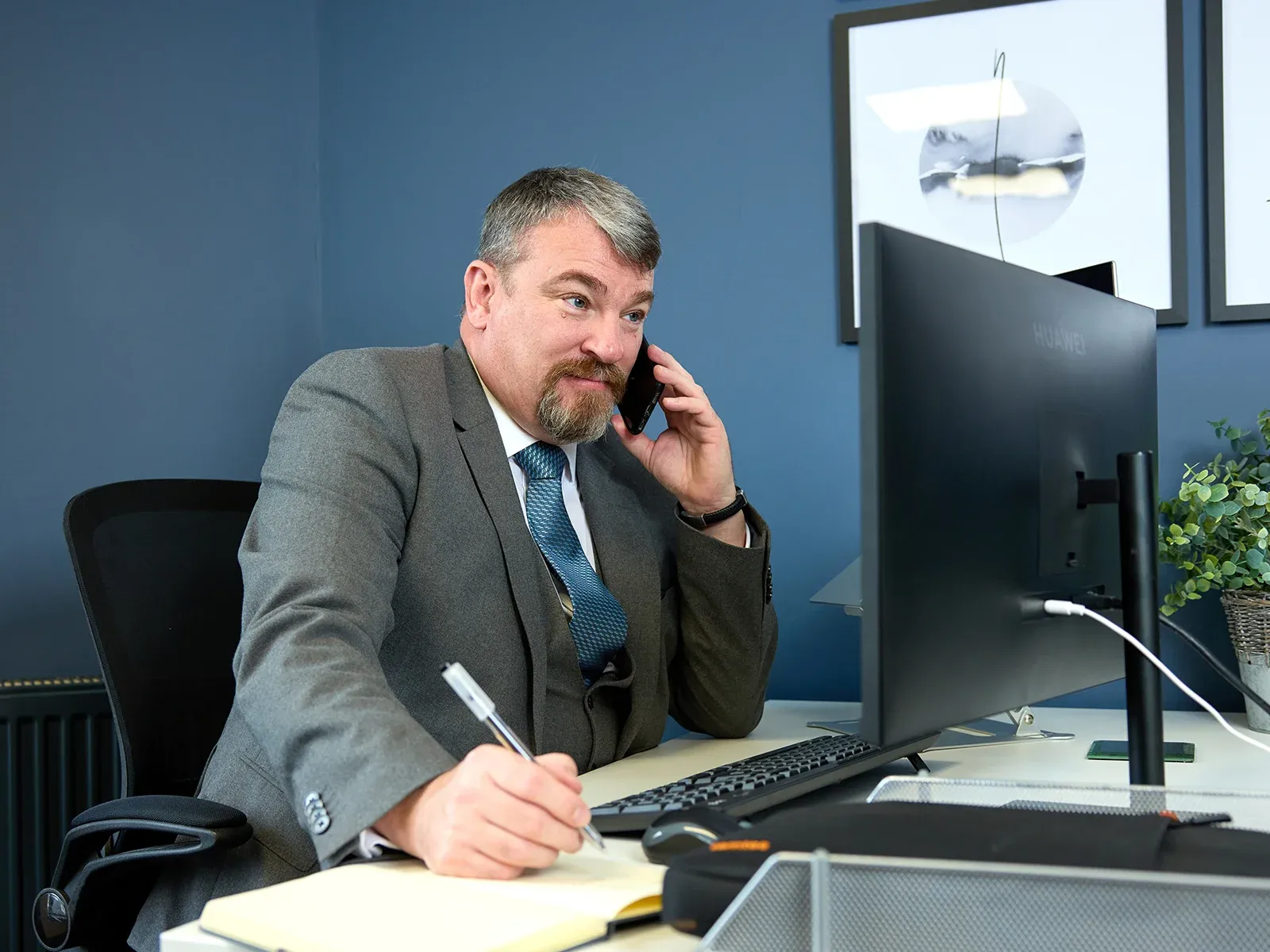 Man in a suit talking on the phone while writing notes at his desk.