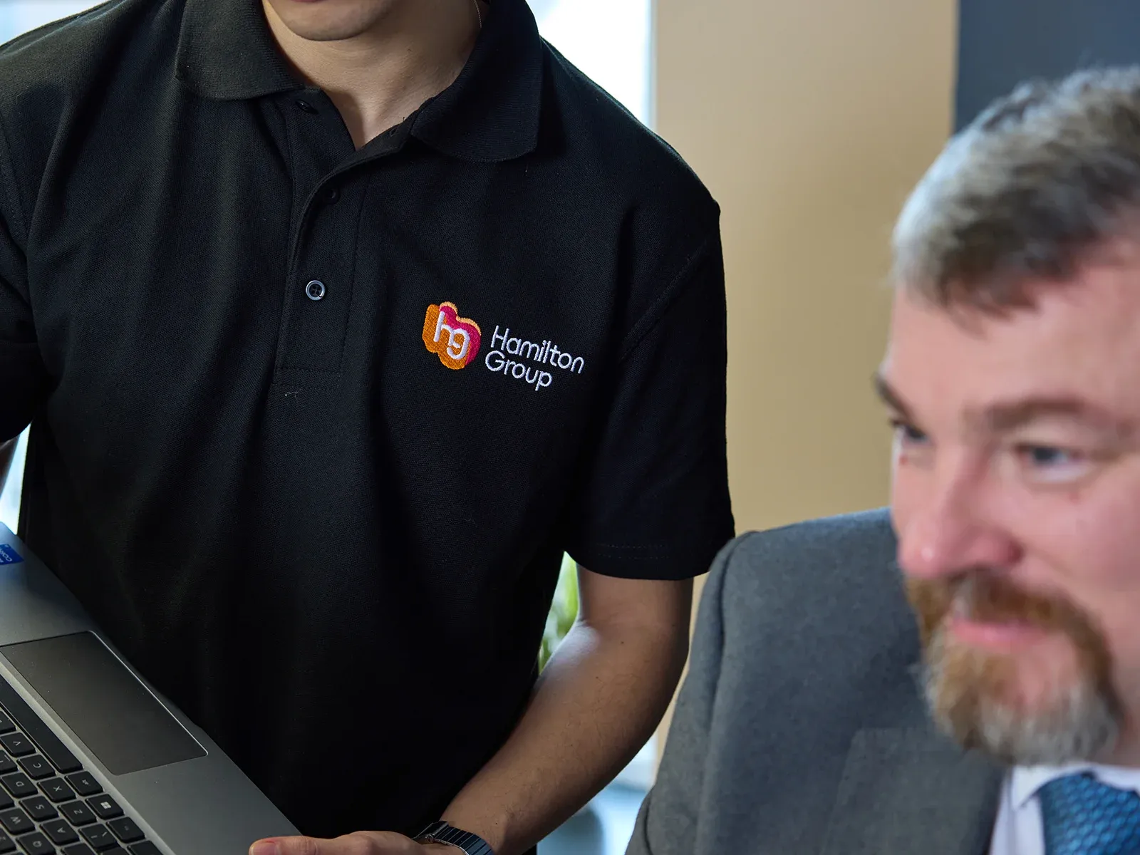 Close-up of a person holding a laptop, wearing a black Hamilton Group polo shirt.