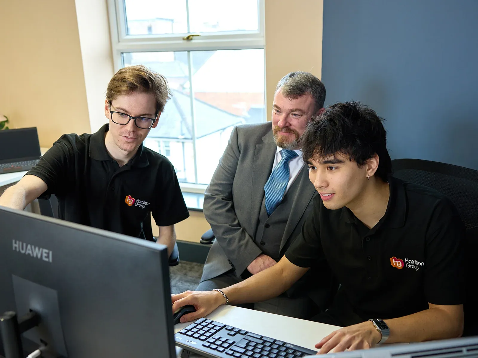 Team collaborating at a computer, with two people in Hamilton Group polo shirts and one in a suit.
