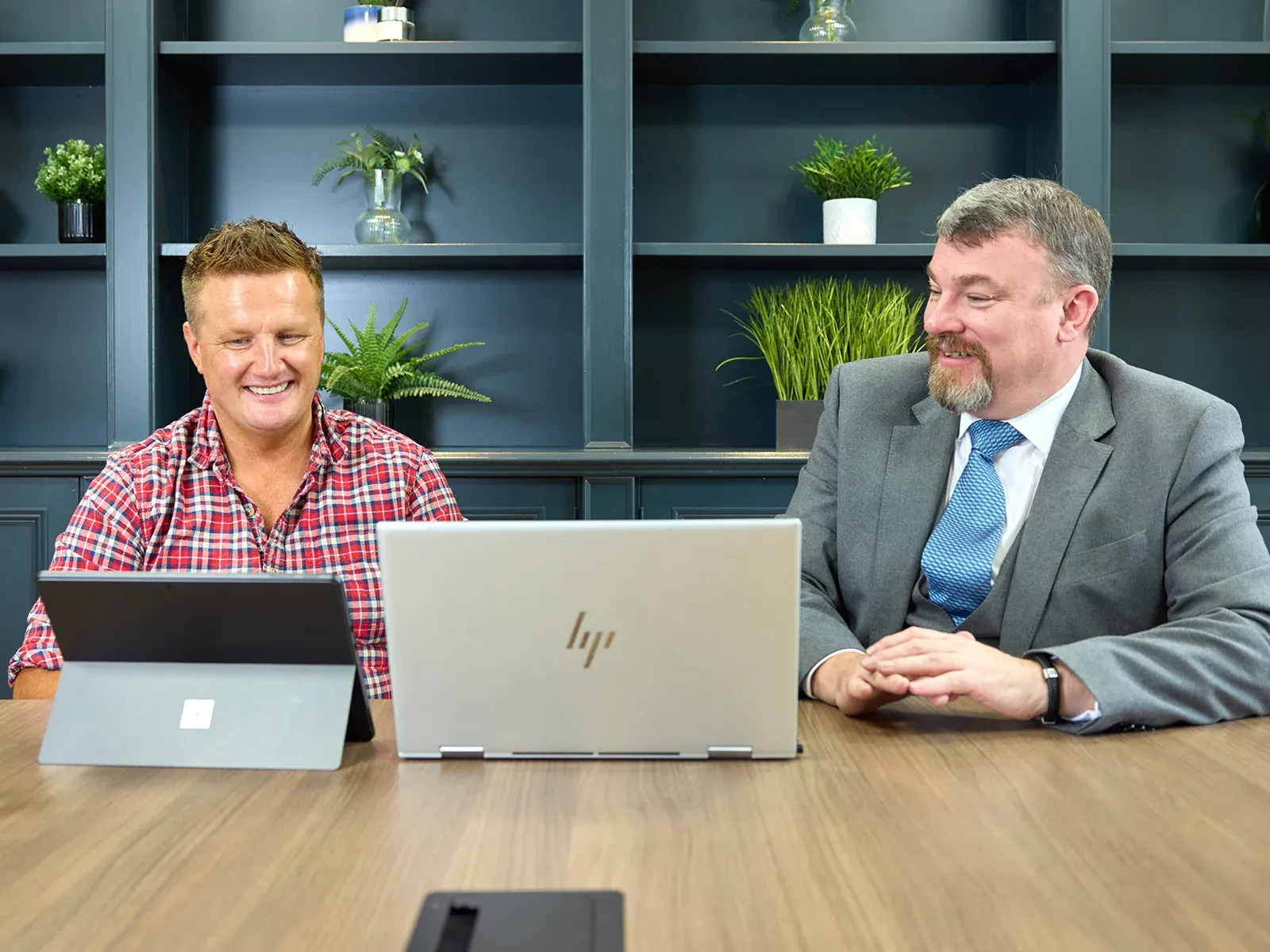 Two men smiling and working together at a conference table with laptops.