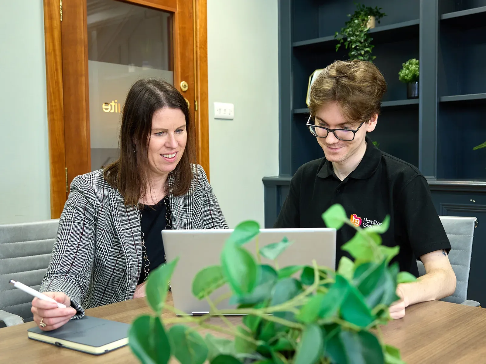 Two colleagues collaborating at a laptop, with a plant in the foreground.