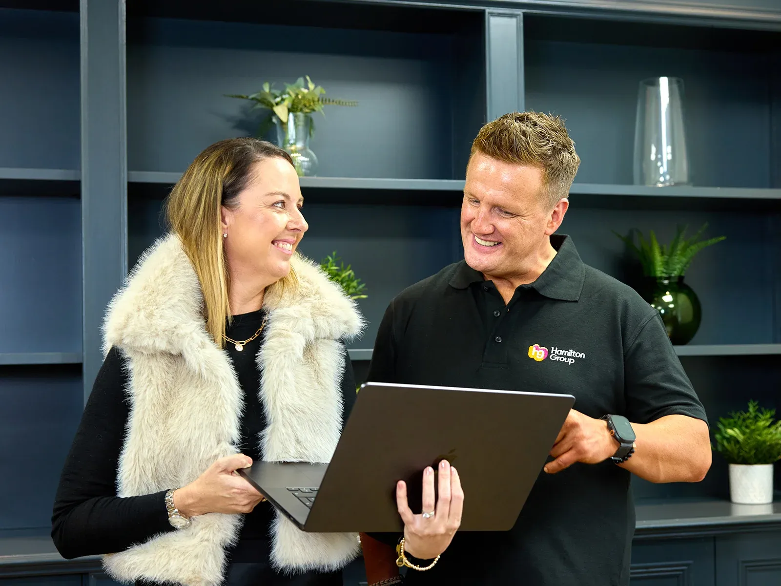 Two colleagues standing and smiling while discussing something on a laptop.