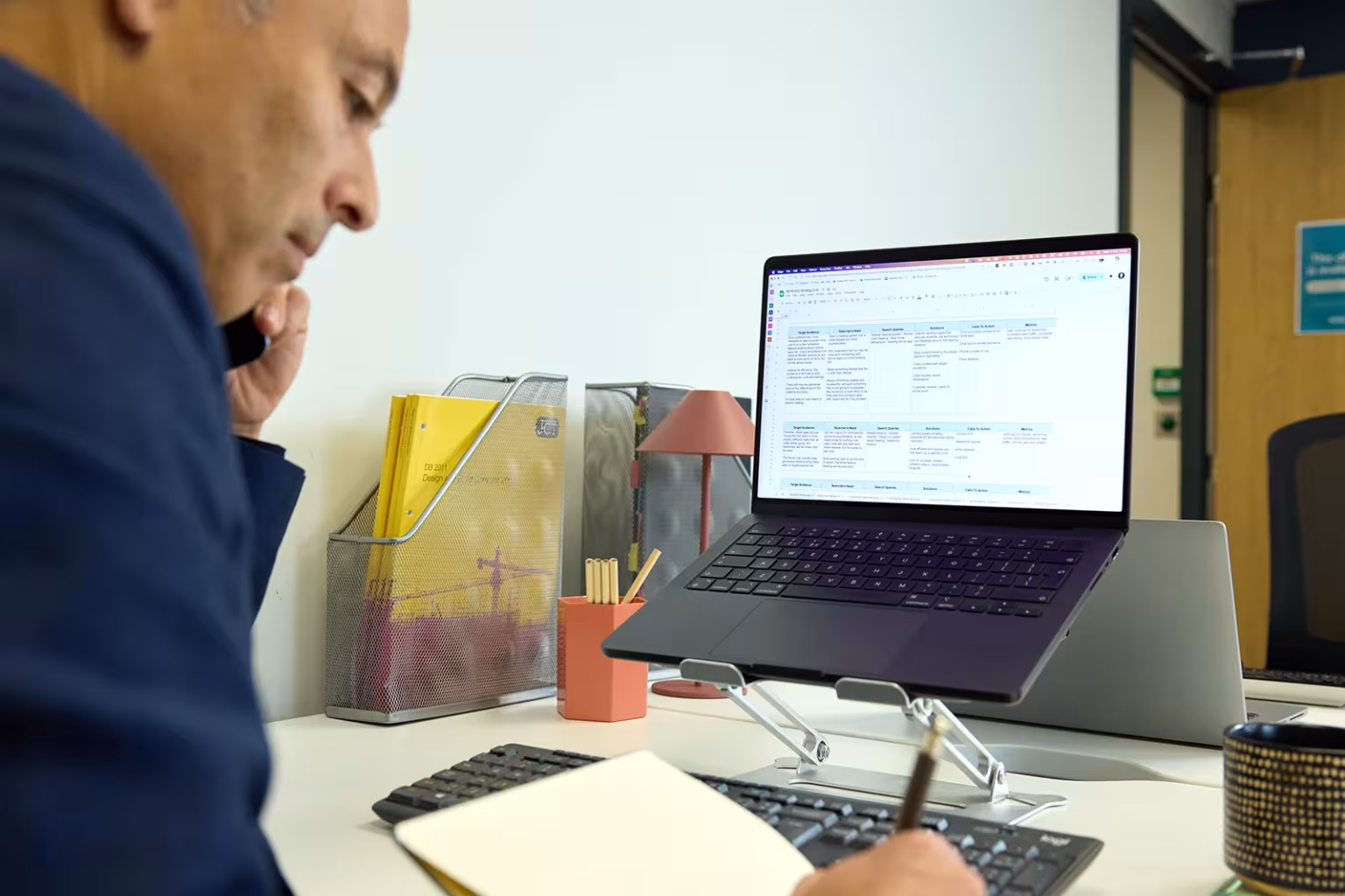 Person on a phone call while taking notes at a desk with a laptop on a stand.