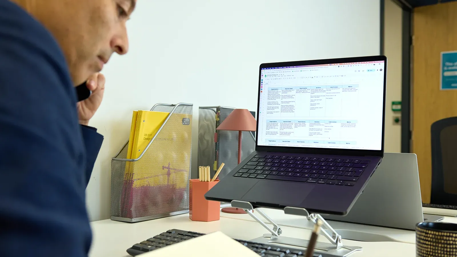 Person on a phone call while taking notes at a desk with a laptop on a stand.