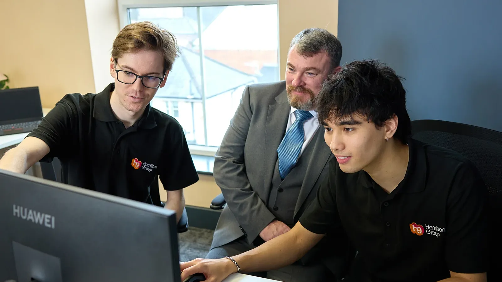 Team collaborating at a computer, with two people in Hamilton Group polo shirts and one in a suit.