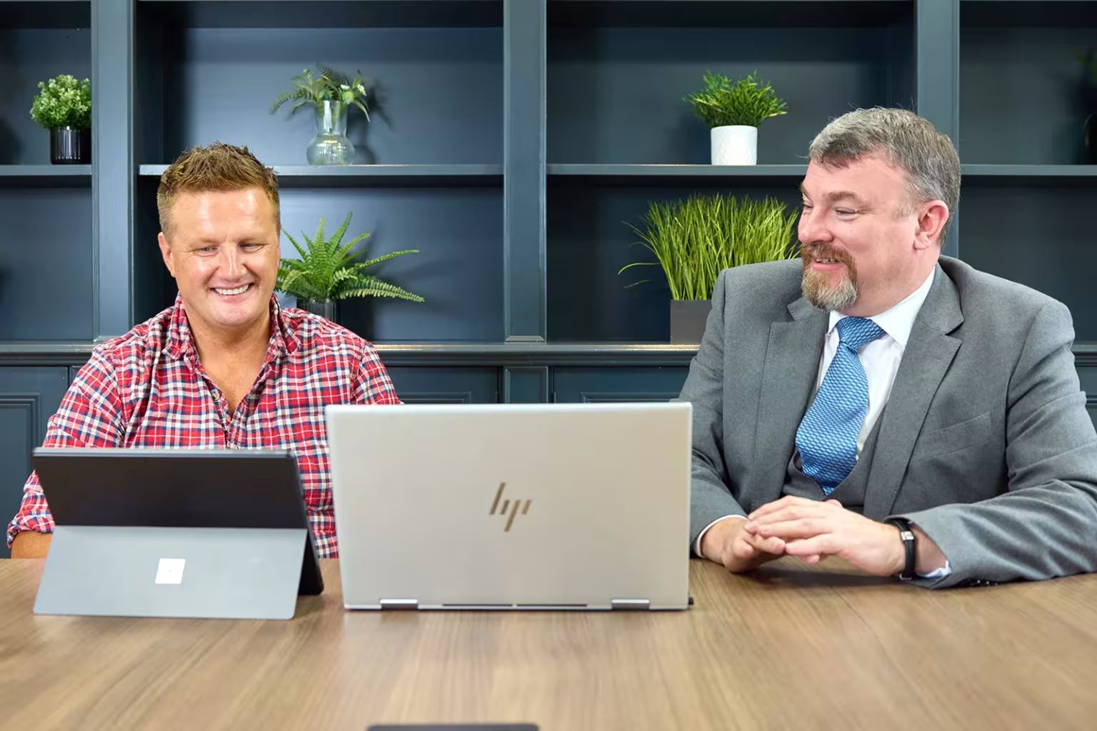 Two men smiling and working together at a conference table with laptops.