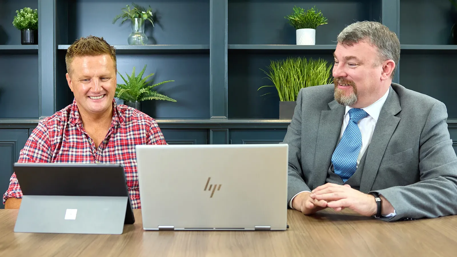 Two men smiling and working together at a conference table with laptops.