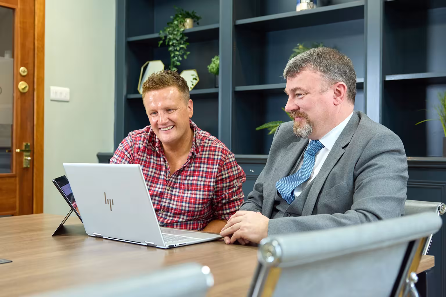 Two men reviewing something on a laptop at a meeting table, smiling during discussion.