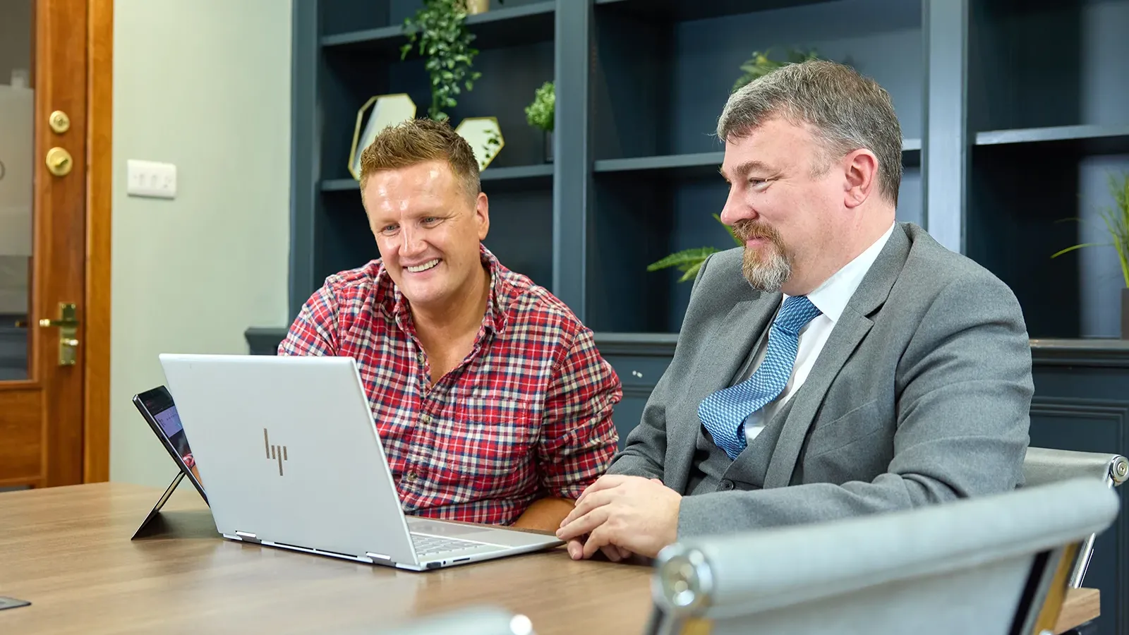 Two men reviewing something on a laptop at a meeting table, smiling during discussion.