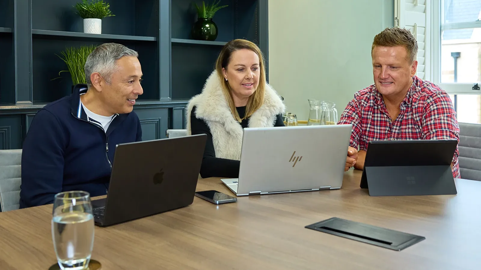 Three colleagues collaborating at a meeting table with laptops open.