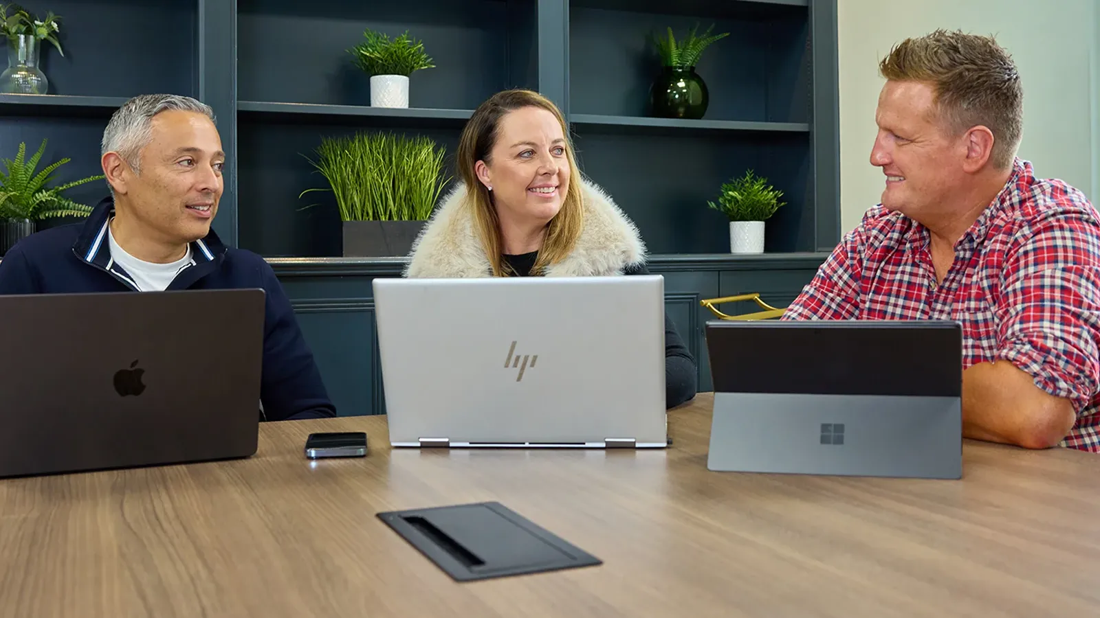 Three people smiling and talking together during a meeting with laptops.
