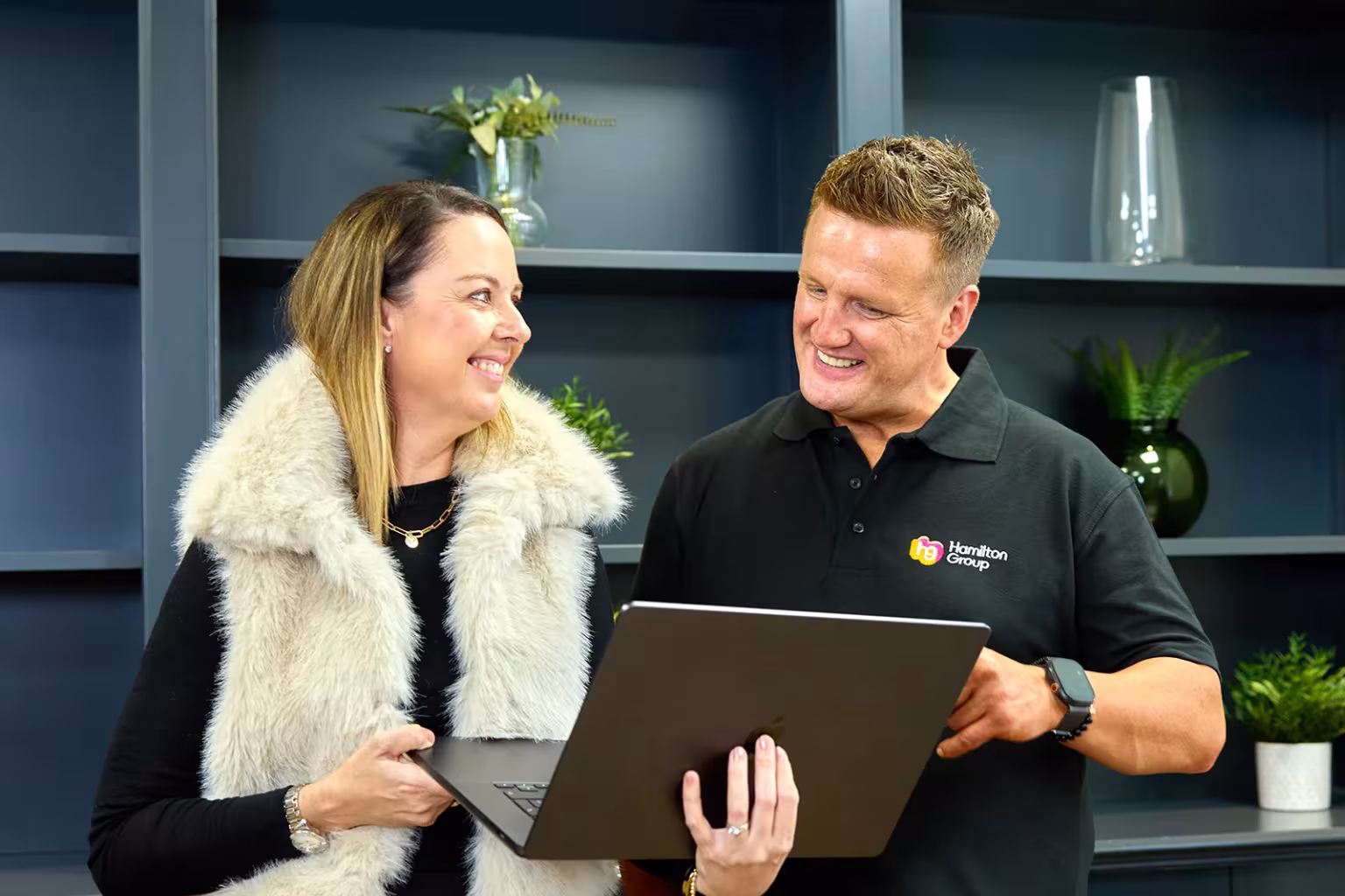Two colleagues standing and smiling while discussing something on a laptop.
