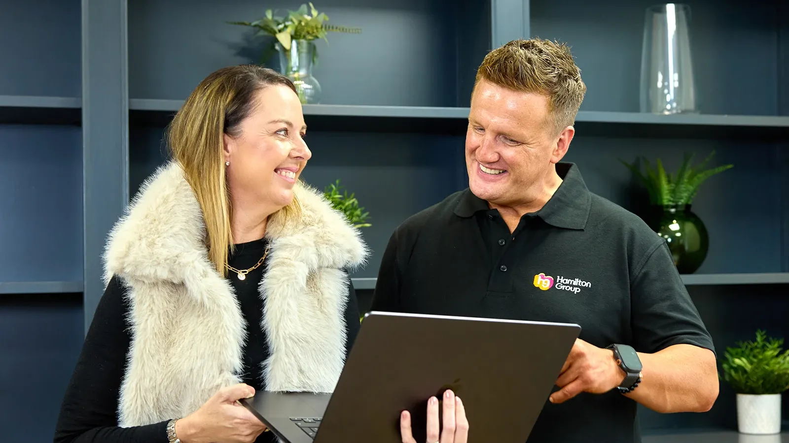 Two colleagues standing and smiling while discussing something on a laptop.